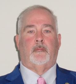 Man with white hair, goatee, wearing blue suit, white shirt, pink tie in front of neutral background