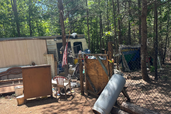 Tan mobile home with lots of debris in yard in a wooded area