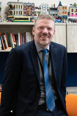 Man in a dark suit and blue tie is smiling and leaning slightly against a desk. Behind, there are shelves with books and Lego models of various buildings.