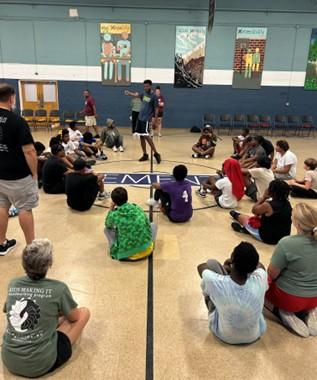 Group of kids sitting on floor of a gym listening to an adult speaker