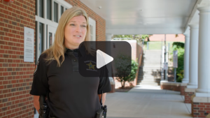 Female in a black shirt with sheriff emblem standing outside a brick building. Play sign in foreground. 