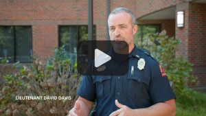 Male in police uniform standing outside a building. Play button in foreground.