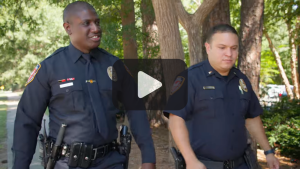 Two males in police uniforms walking together. Play button in foreground.