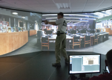 Male standing in middle of a room in front of a large screen displaying what appears to be a library. Man has a gun drawn.