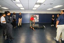 Group of people in a room with mats placed on floor and walls. A person lays on the floor in the middle with an instructor teaching a technique on them.