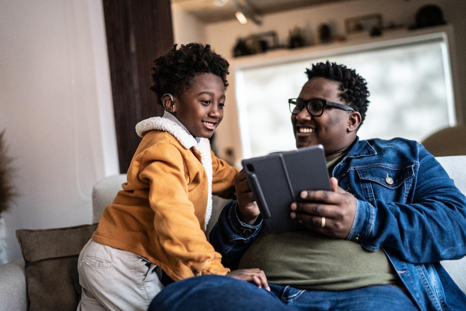 A father smiles and shares content on a tablet device with his young son who wears a hearing aid.