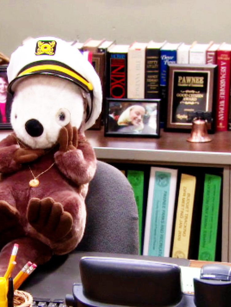 A stuffed toy otter wears a ship captain's hat and sits in an office chair from a scene in the Parks and Recreation television show.