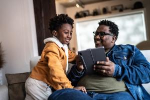 A father smiles and shares content on a tablet device with his young son who wears a hearing aid.