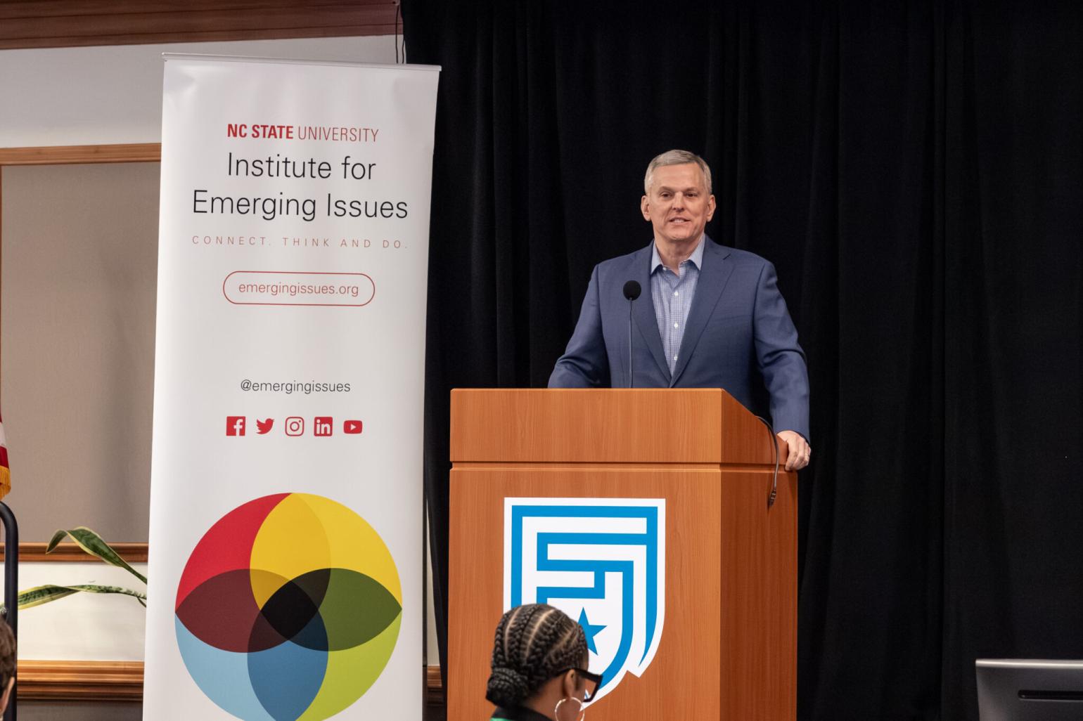 Leader in a gray suit addresses conference attendees from a wooden podium