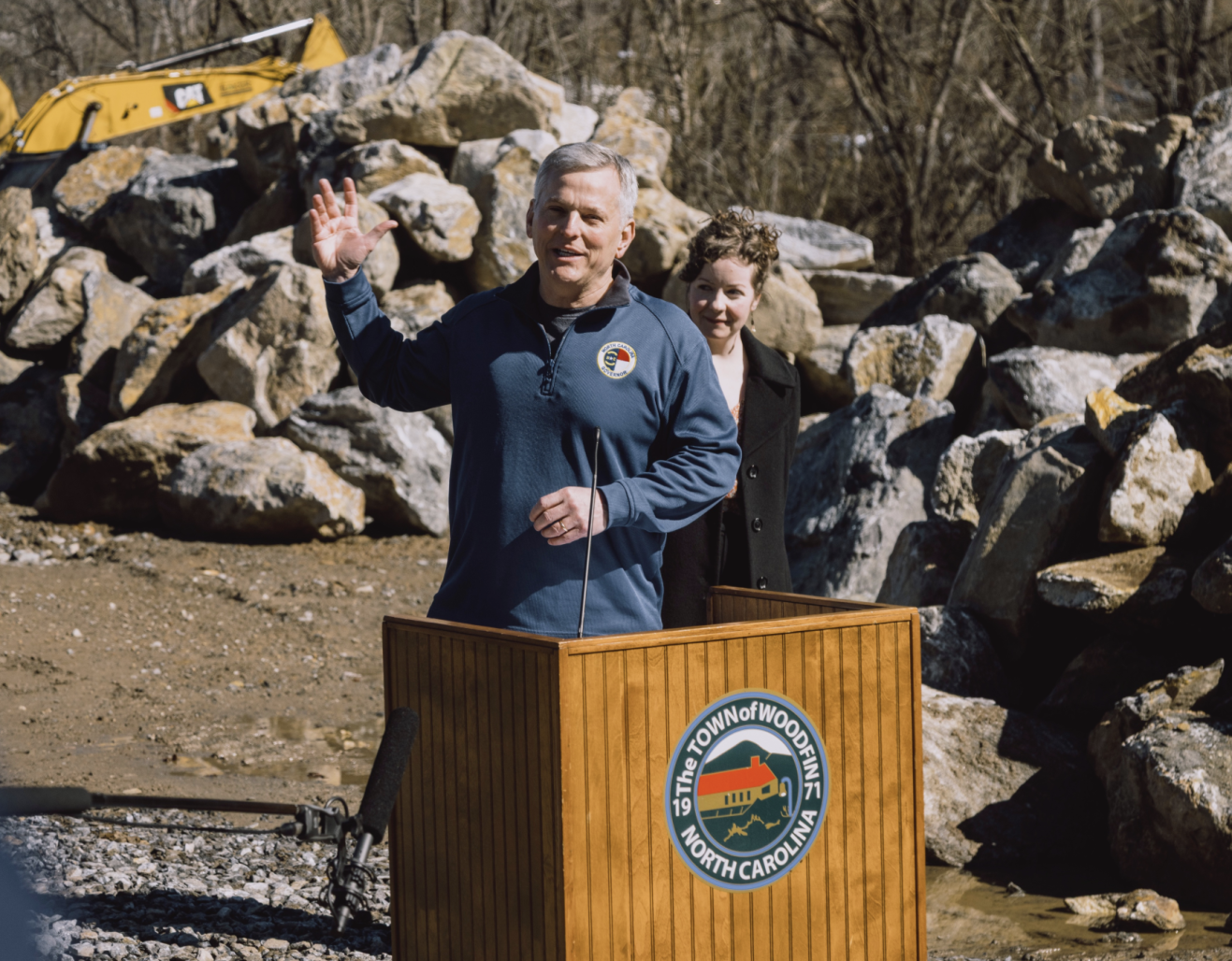 A leader gestures from a podium in an outdoor setting with heavy machinery in the background