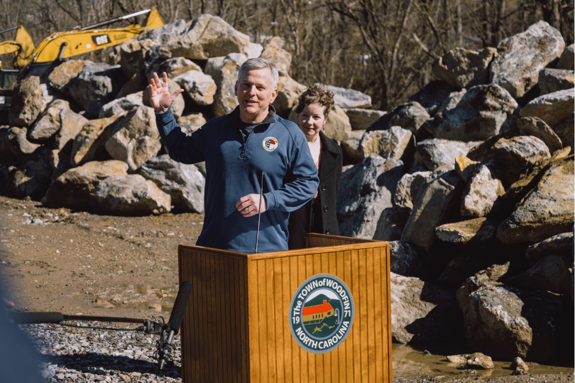 A leader speaks at a wooden podium in an outdoor setting