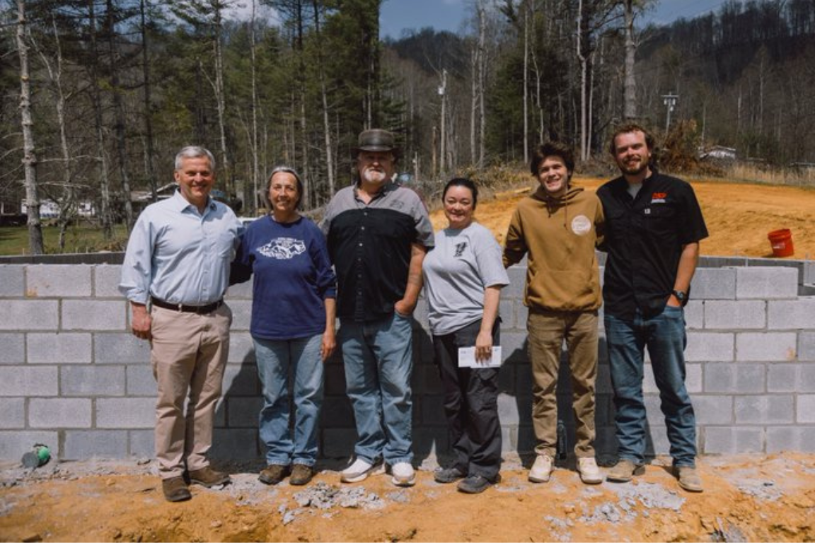 Six people stand in front of the foundation of a home, partially constructed