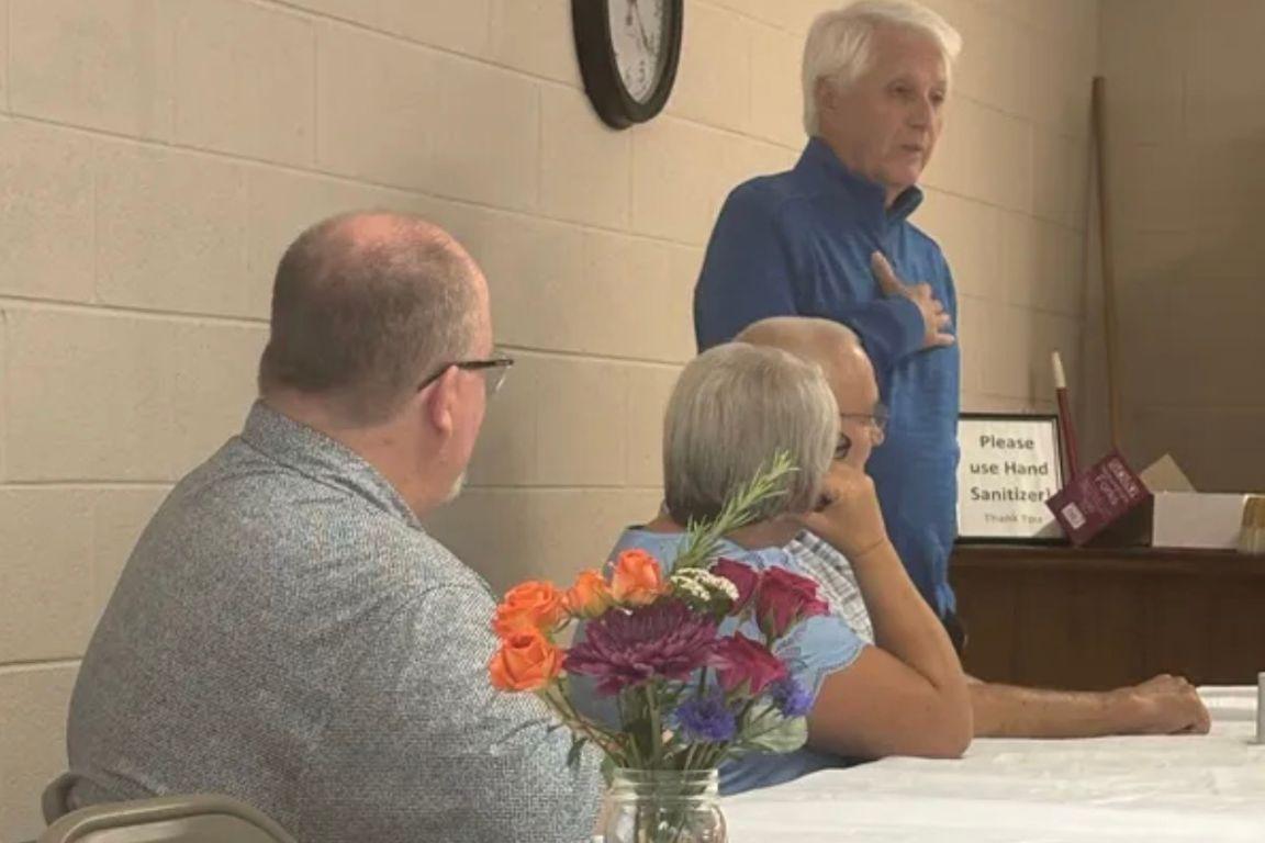 Three people sit at a table together while being addressed by a man who is standing