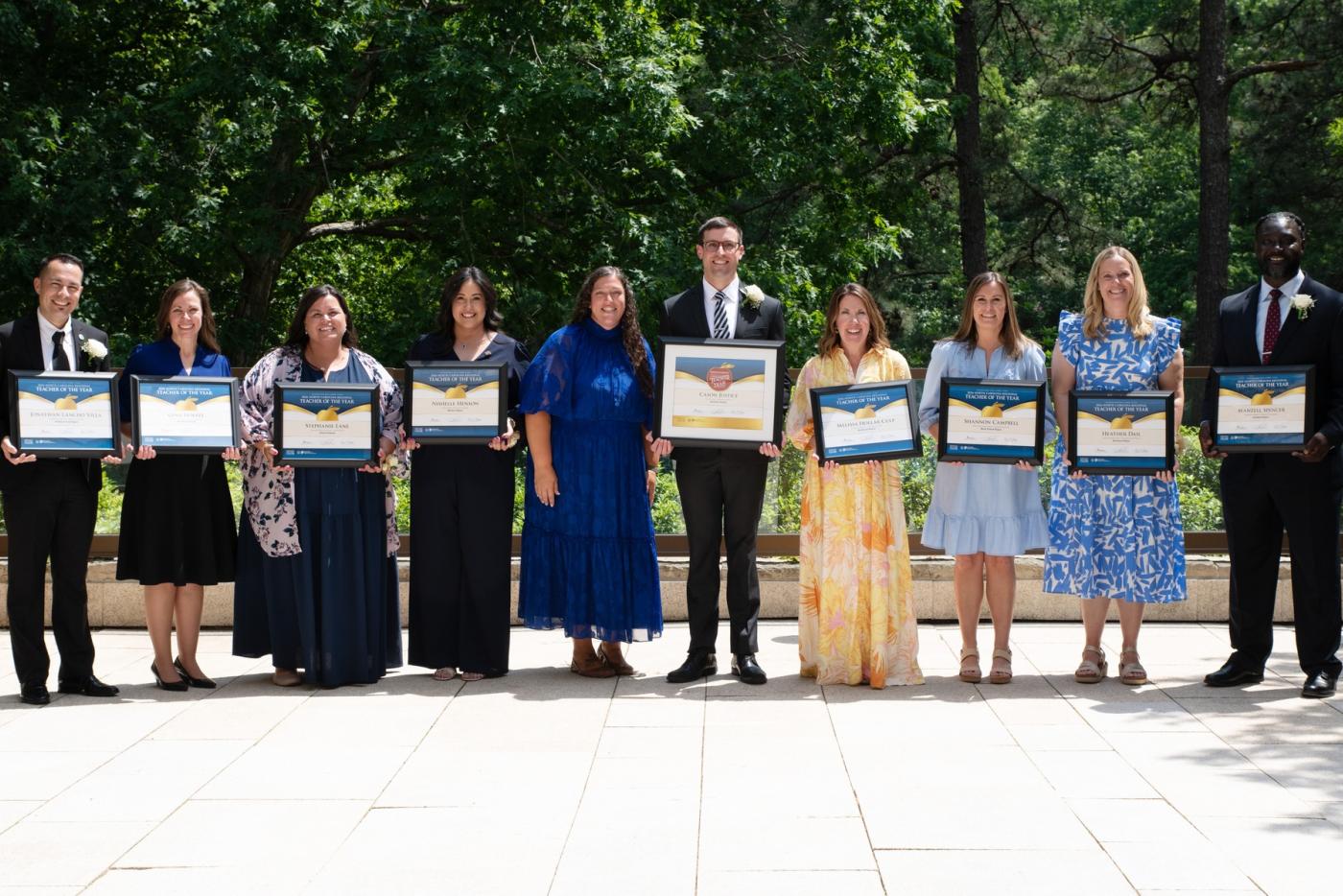 Ten people wearing business attire are holding certificates in an outdoor setting