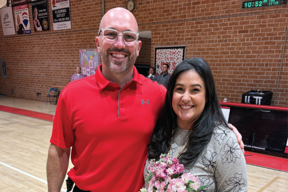 Two teachers pose in a basketball gymnasium with a brick wall background