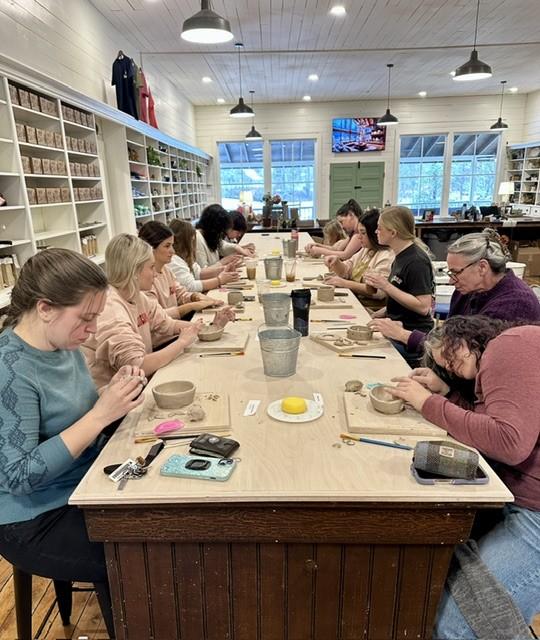 Students sit along a craft table holding pottery projects and paint