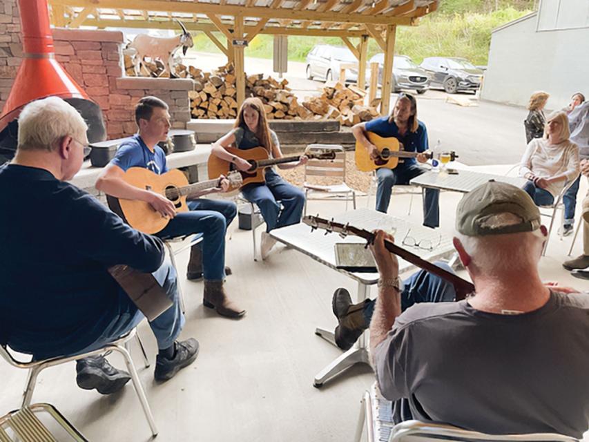 Six adults sit in a circle holding acoustic guitars in an outdoor setting