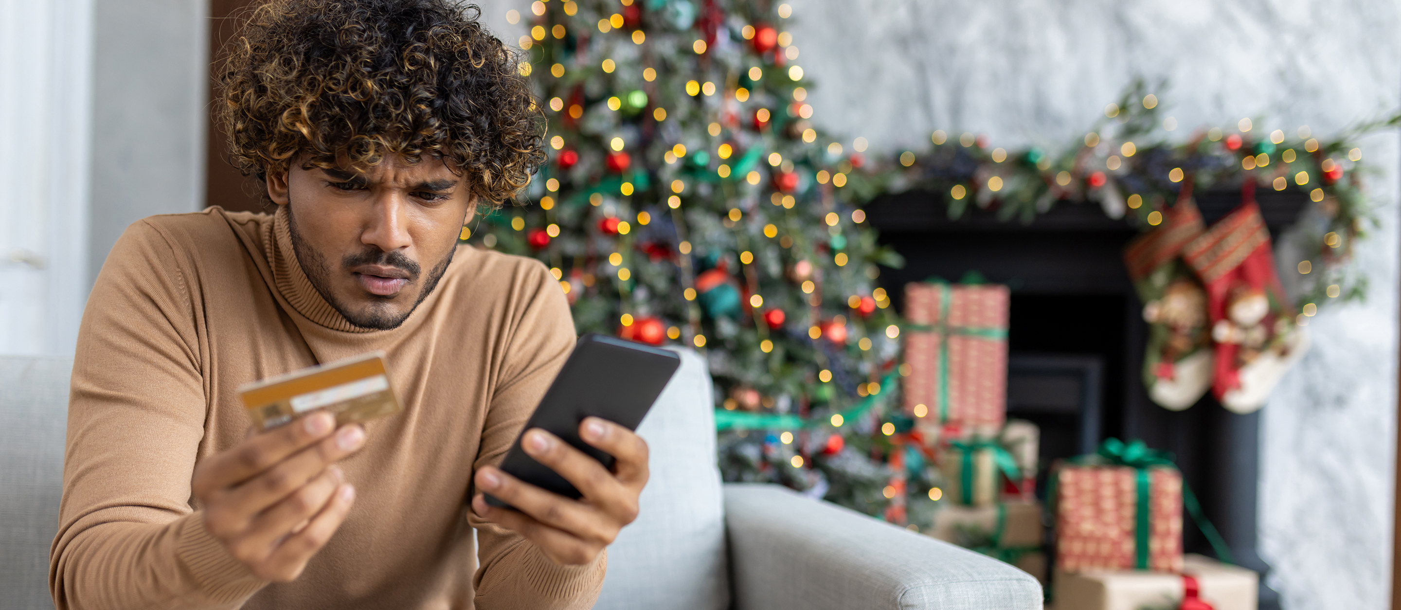 A young, Hispanic man frowning while holding a credit card and phone while seated on a couch in a room decorated with a Christmas tree.