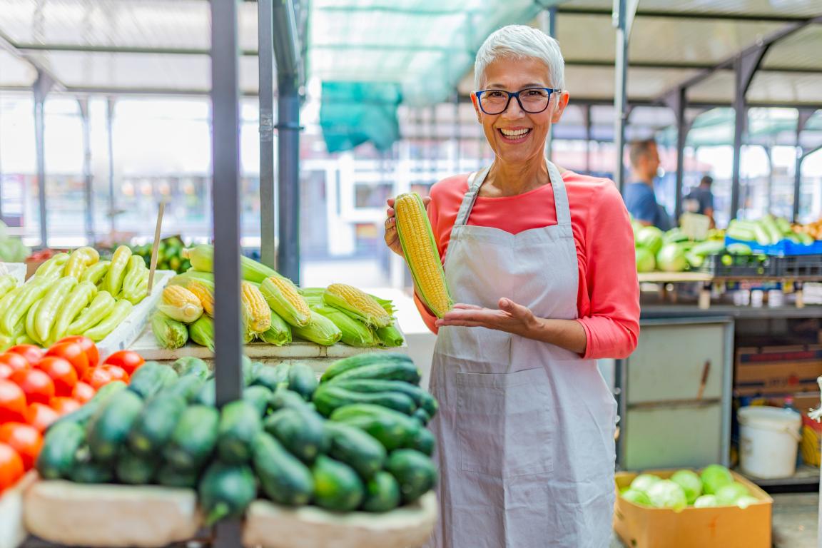 Smiling, gray-haired women wearing an apron holding an ear of corn in a vegetable produce market.