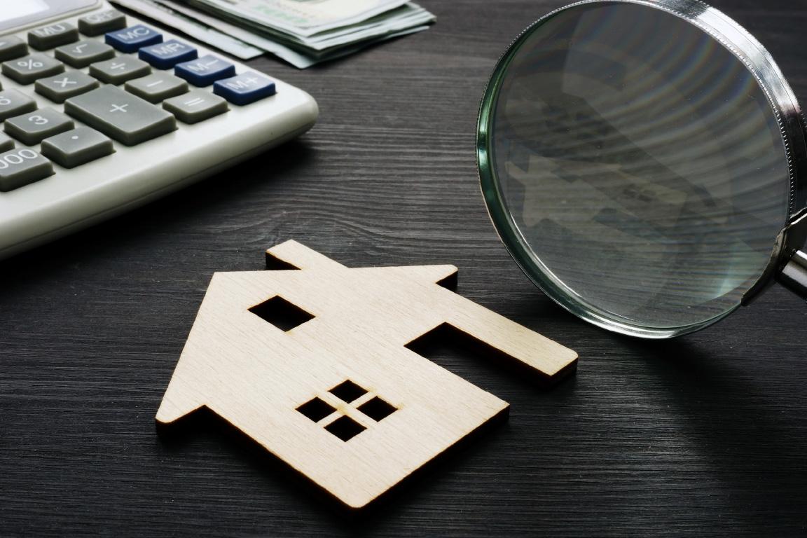 A magnifying glass next to a small, flat, wooden cut-out of a house lying on the top of an office desk.