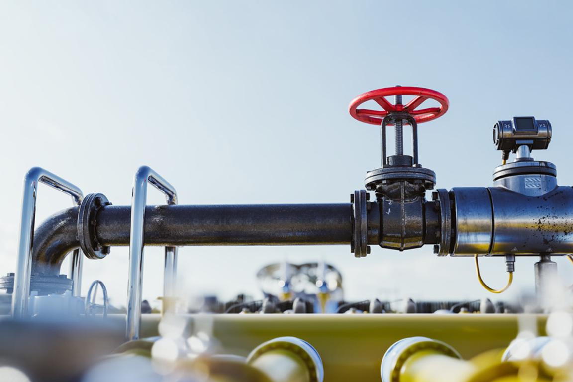 A close-up of an industrial pipeline with a black pipe featuring a red valve wheel. The background shows a clear blue sky, conveying a sense of functionality and precision.