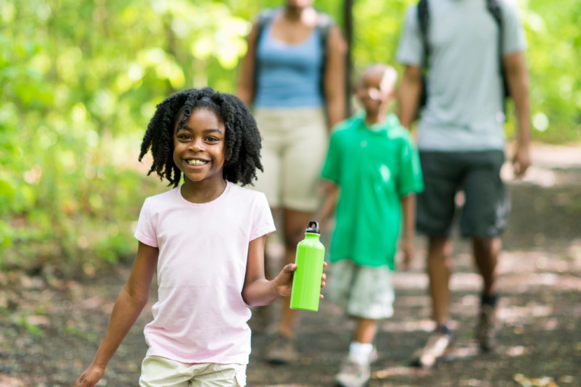 A young girl, smiling walks with her family outside on a sunny day in the park.