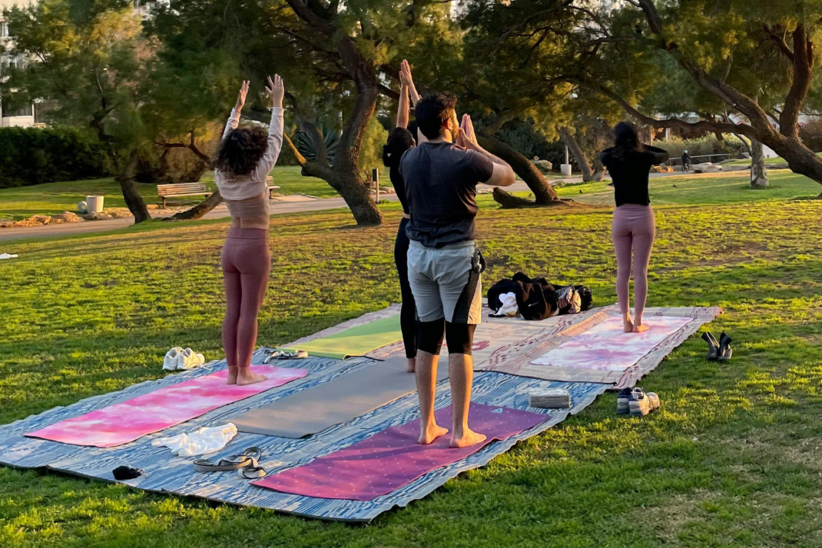 A group of people practicing yoga on mats in a sunny park, surrounded by trees and green grass.