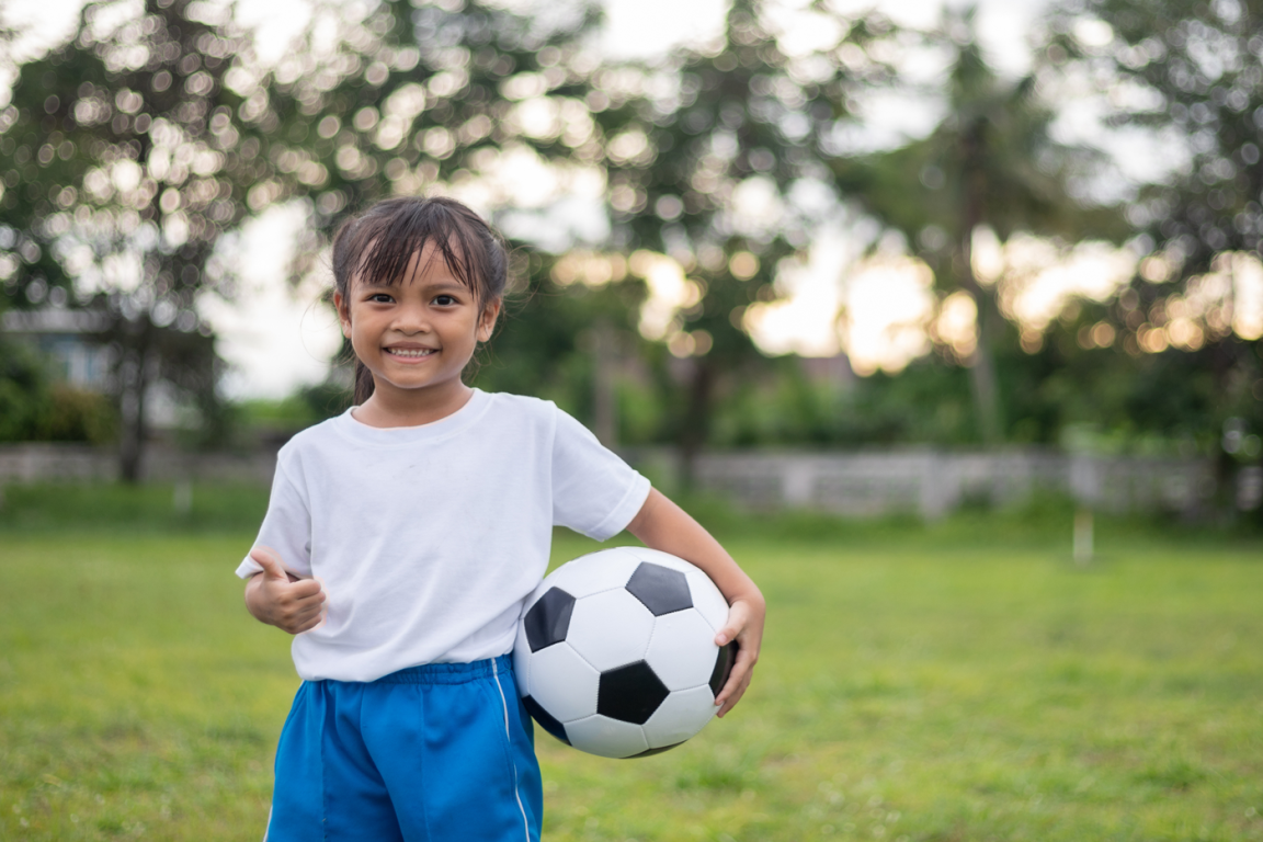 A young, smiling girl holding a soccer ball stands outside on a grassy field surrounded by trees.
