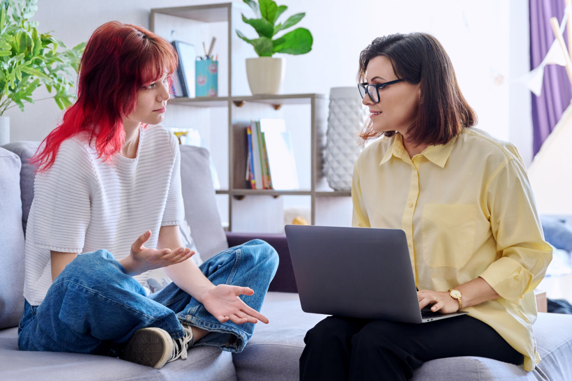 A female social worker with a laptop and a young teenage girl are seated on a couch talking.