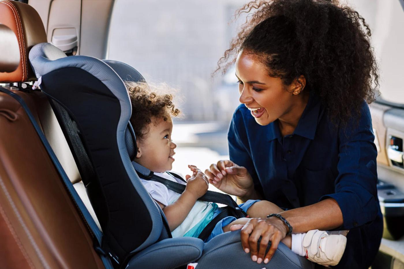 Smiling mother buckling her young toddler in a child car seat.