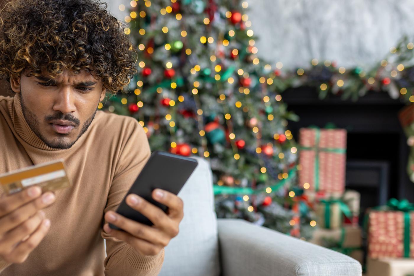 A young, Hispanic man frowning while holding a credit card and phone while seated on a couch in a room decorated with a Christmas tree.