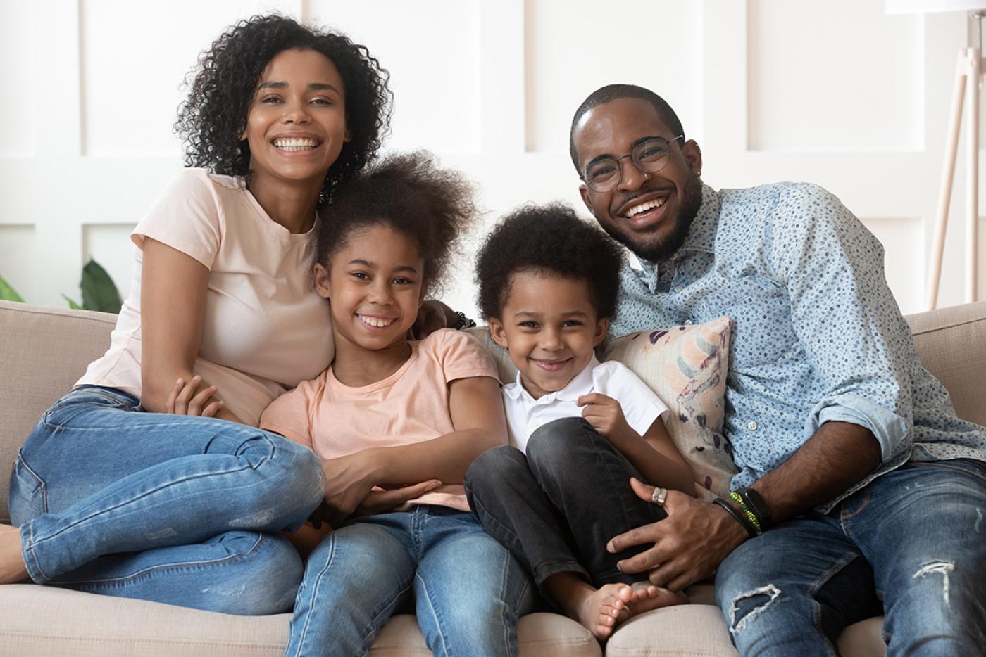 A smiling family of a mother, father, and two young children sitting closely together on a couch.
