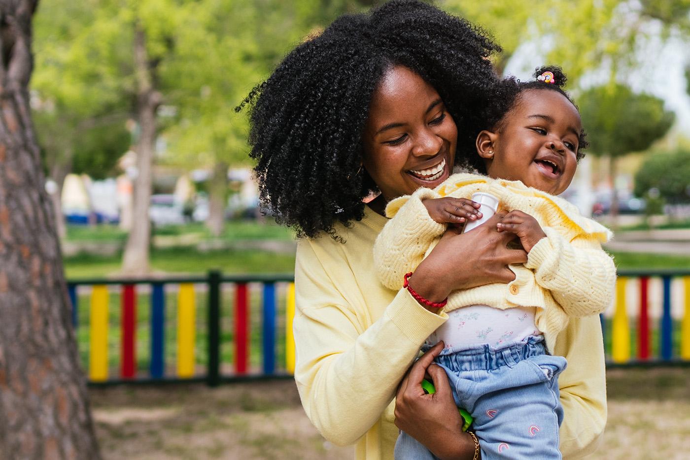 A young, black mother holding her infant child in a park as they both smile.