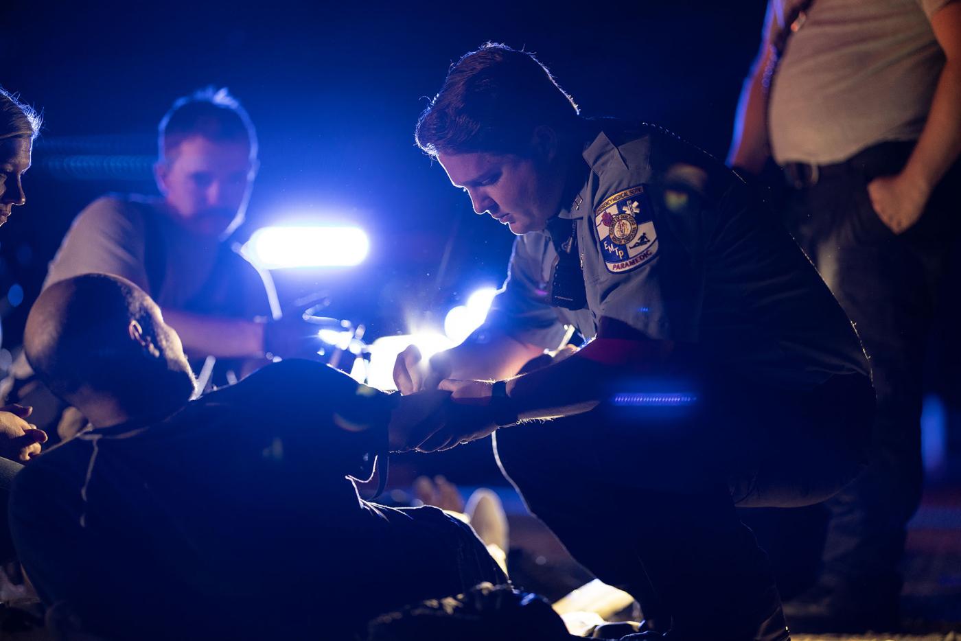 An emergency medical technician tends to an injured person on the ground at night, illuminated by emergency lights. 