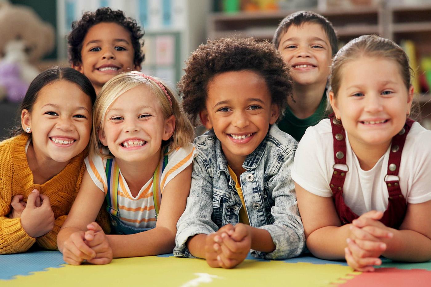 Group of smile kindergartener children next to each other resting their elbows on the padded classroom floor.