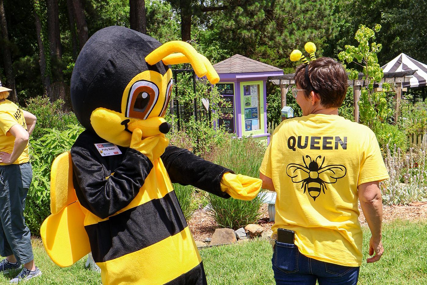 A person in a bee costume playfully interacts with someone wearing a "Queen" bee-themed shirt. They stand in a lush, green garden, evoking a cheerful atmosphere.