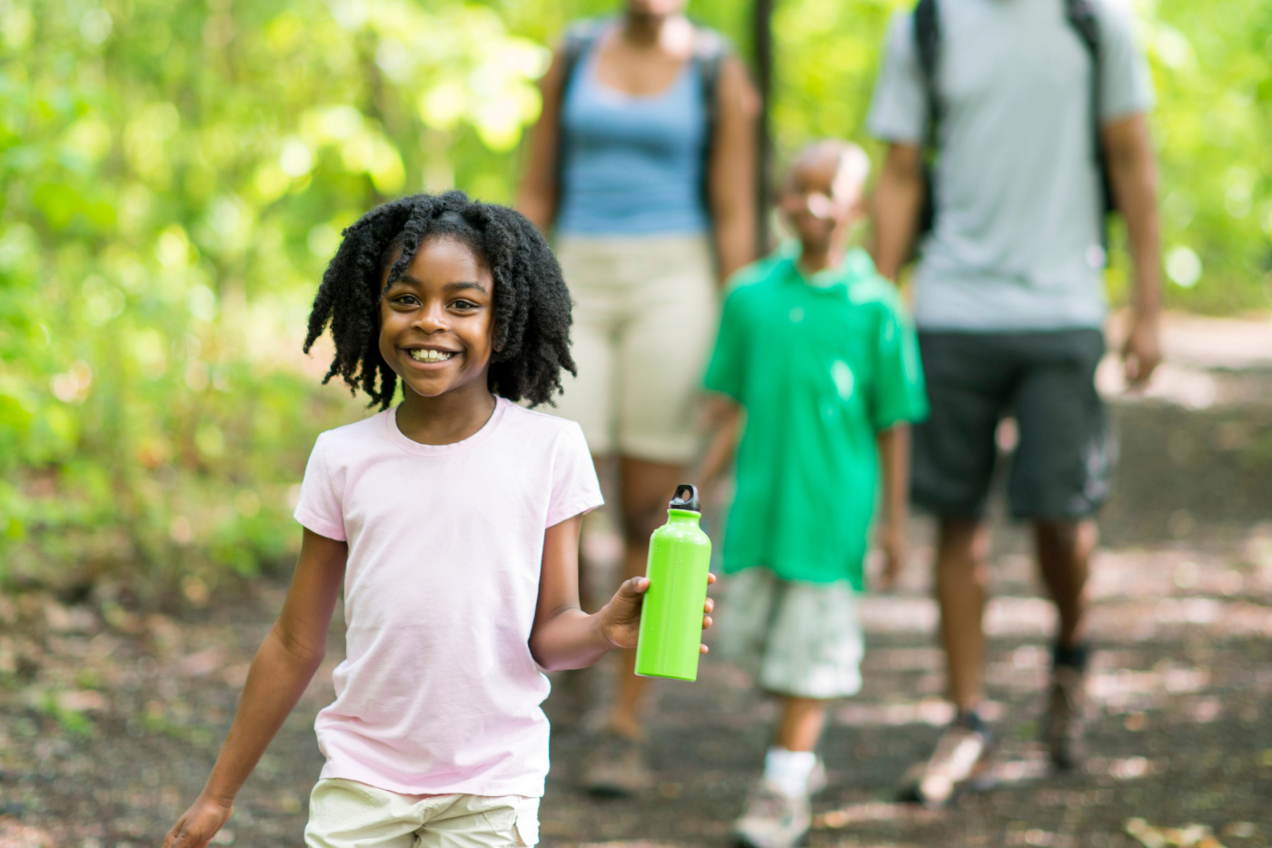 A young girl, smiling walks with her family outside on a sunny day in the park.
