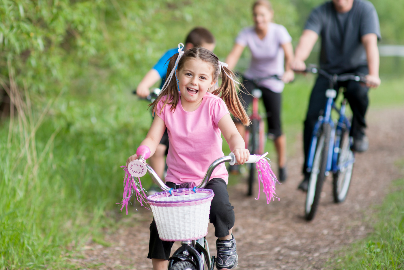 A young girl on a bicycle, smiling as she rides with her family on a park trail on a sunny day.