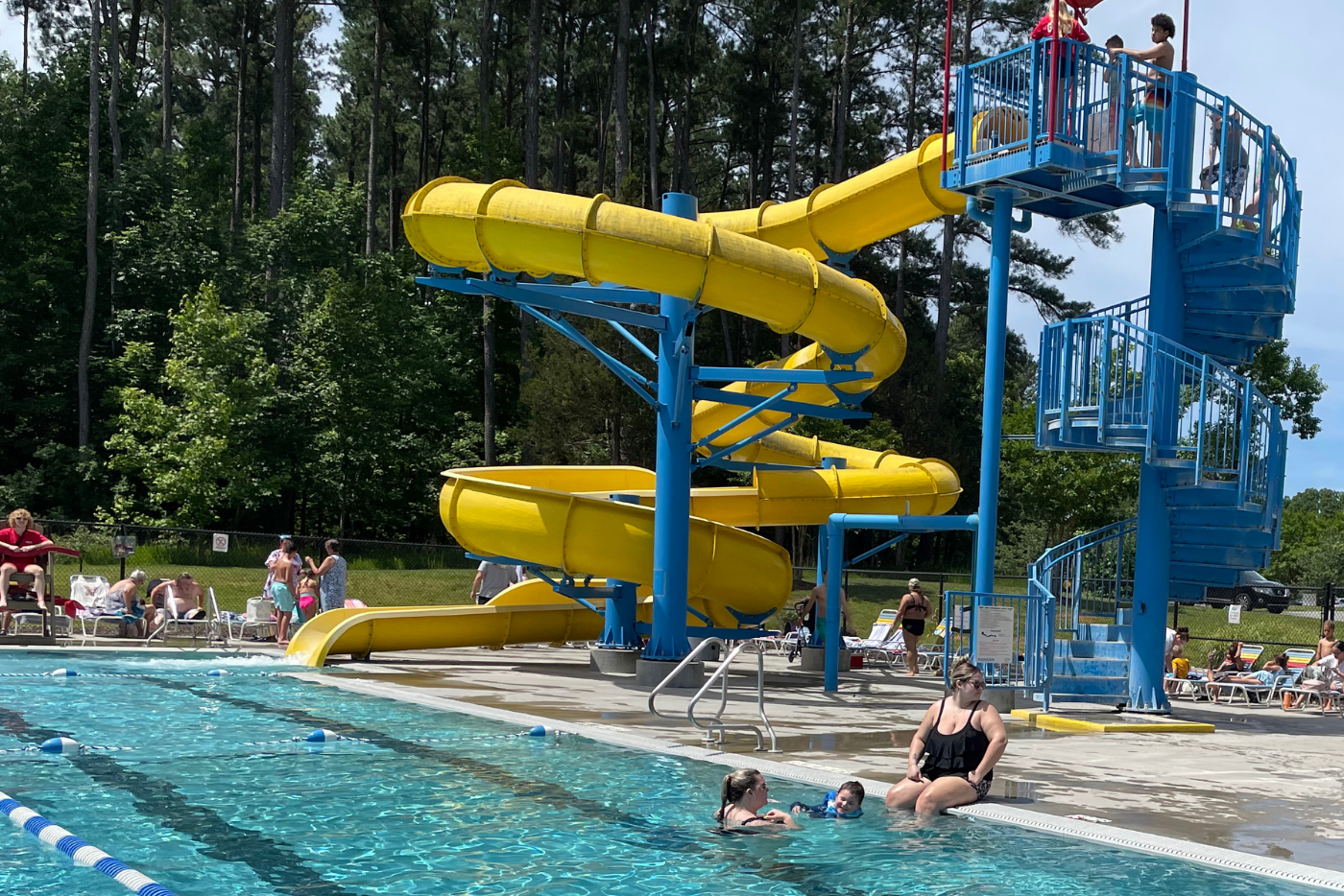 People enjoying a sunny day at the Northeast Park aquatic center that includes a large, yellow twisty pool slide.