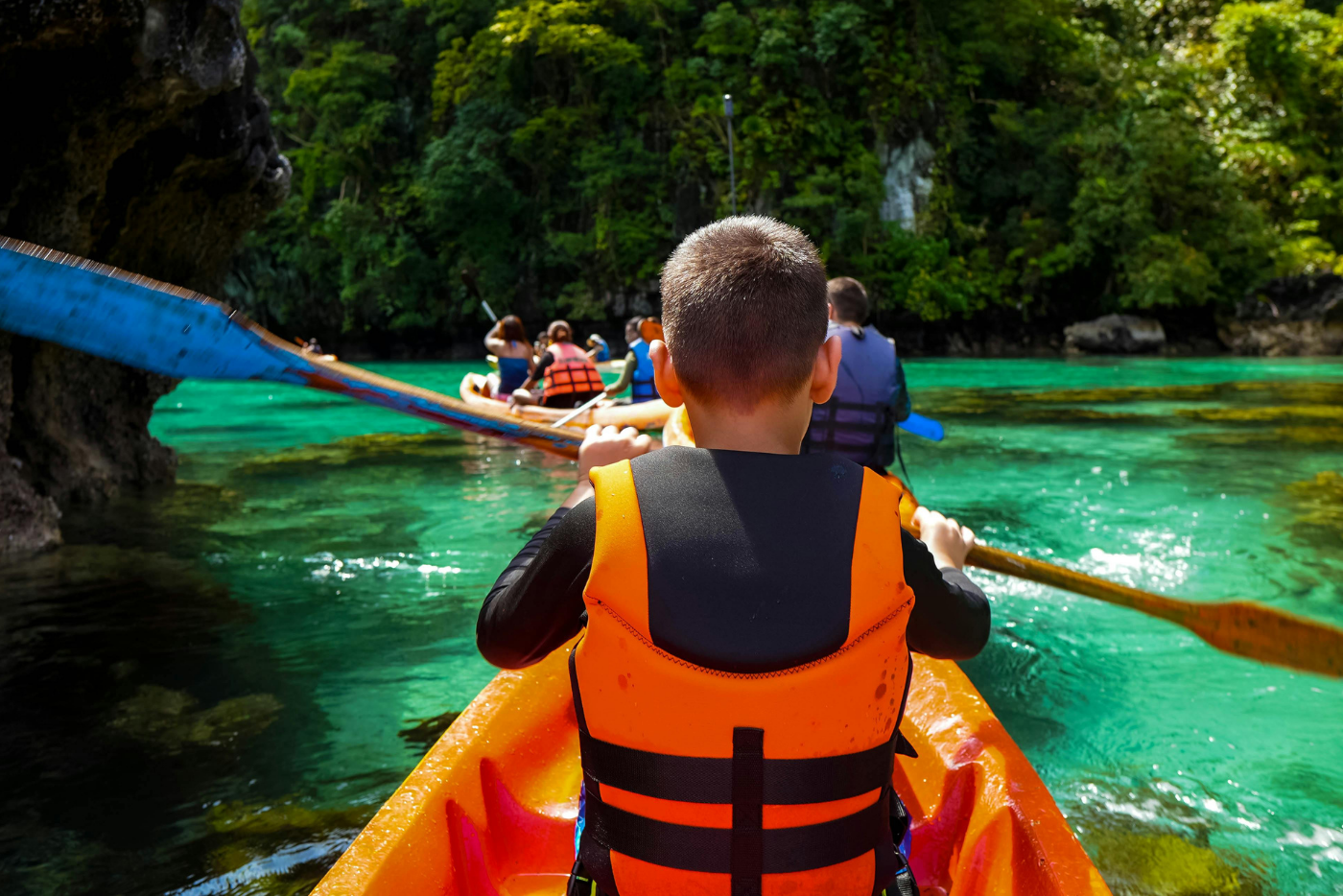 A boy wearing an orange life jacket paddles a canoe on calm water, enjoying a sunny day outdoors.