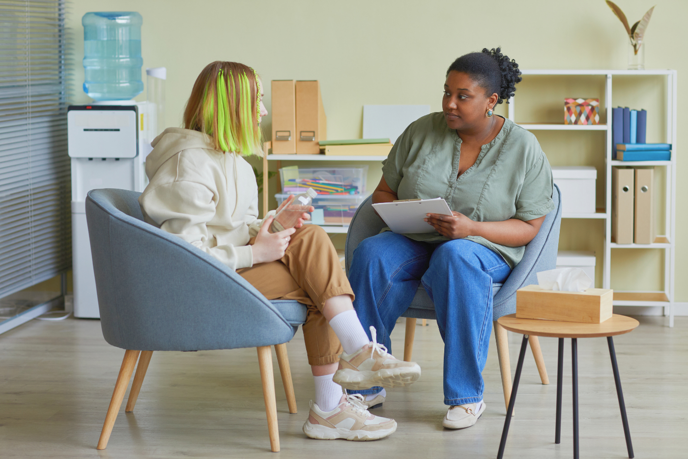 A female social worker seated in a chair with a clipboard speaks with a female teenager seated across from her.