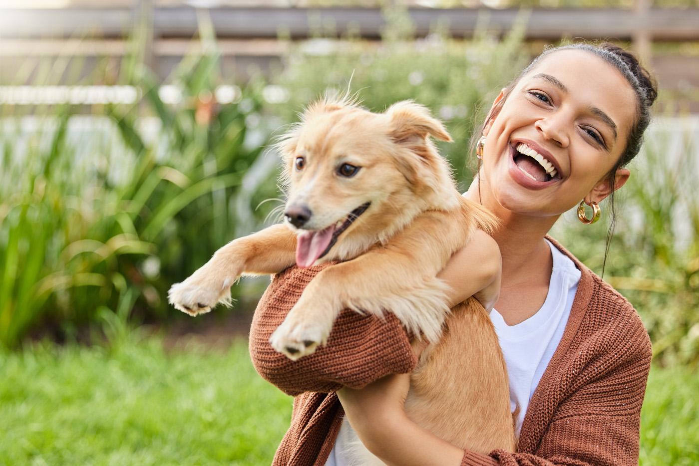 A young smiling woman holding her medium sized, tan colored dog outside in the grass on a sunny day.