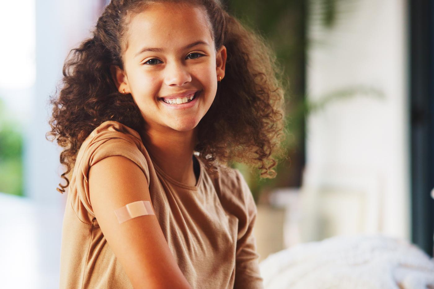 A young, smiling girl with her sleeve rolled up revealing the bandage received after getting her seasonal flu vaccine.