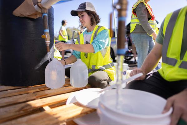 A young woman wearing a yellow vest dispensing water into gallon jugs.