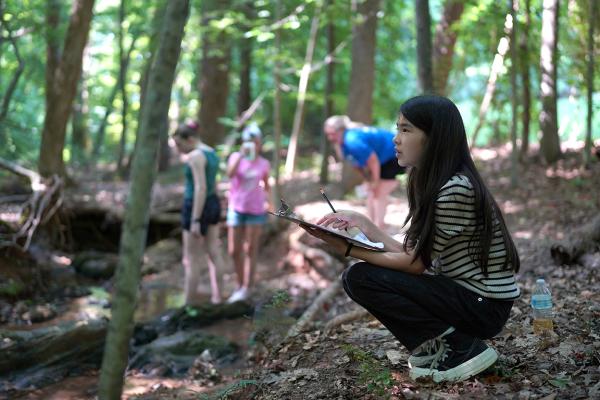 A young girl with clipboard making nature observations in a wooded area with other youth exploring the surrondings.