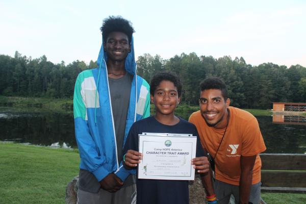 Three smiling boys stand outdoors near a lake, with the middle boy holding a Character Trait Award certificate from Camp HOPE America.