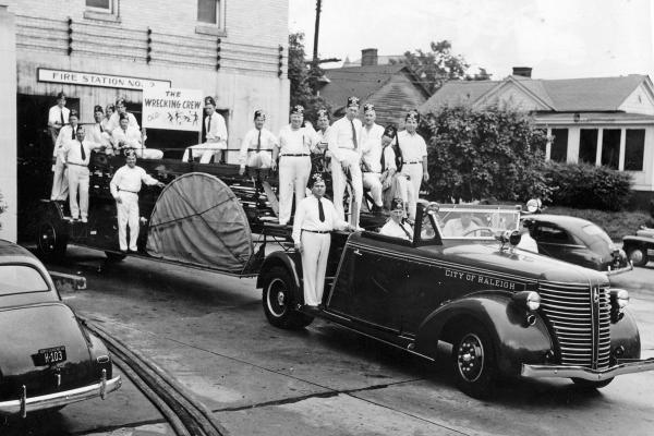 A black and white photo of 17 men with matching outfits standing on a historical fire truck parked at a brick building. 