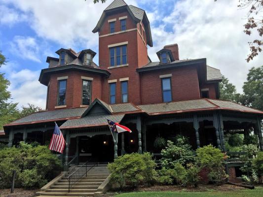 An asymmetrical four story brick building with a slate roof with concrete stairs leading to a wraparound porch