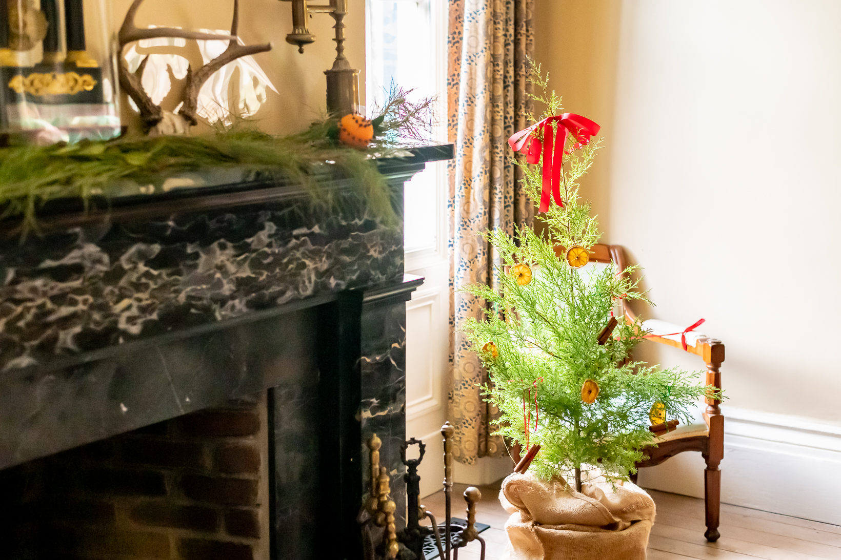 Small Christmas tree decorated with dried oranges and red ribbon. To the left there are Christmas decorations on the fireplace mantle. 