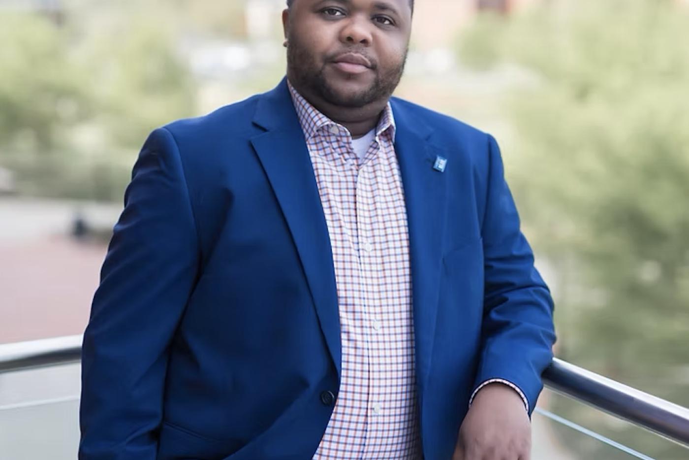 a Black man in a blue suit leans against a railing